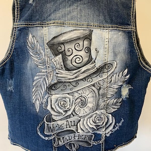 May include: A distressed denim vest with a hand-painted design featuring a Mad Hatter hat, pocket watch, roses, and feathers. The phrase "WE'RE ALL MAD HERE" is displayed on a banner. The artwork is in shades of gray and white.