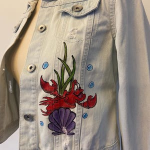 May include: Light blue denim jacket with a distressed design. The jacket features a hand-painted illustration of a red crab, green seaweed, a purple seashell, and blue bubbles. The jacket has silver buttons and two front pockets.