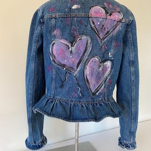 May include: A blue denim jacket featuring three large, hand-painted purple hearts on the back. The hearts have black outlines and white accents. The jacket has a ruffled hem and cuffs, with paint splatters.