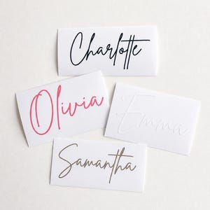 May include: Four white rectangular cards with names in cursive script. The names are "Charlotte" in black, "Olivia" in pink, "Emma" in white, and "Samantha" in gold. The cards are arranged on a white surface.