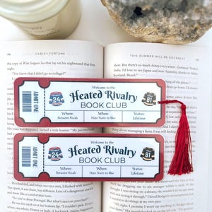 Könnte beinhalten: Zwei ticketförmige Lesezeichen mit dem Text "Heated Rivalry Book Club" und einer roten Quaste. Die Lesezeichen befinden sich auf einem offenen Buch. Die Tickets haben die Worte "Admit One" und die Zahlen 24 und 81.