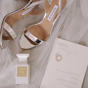 Può includere: Sandali con tacco Aquazzura argentati metallizzati, un flacone di profumo Tom Ford Soleil Blanc, un invito di nozze e un anello d'oro su una superficie bianca. I sandali hanno sottili cinturini e un tacco alto.