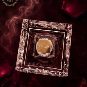 May include: A square glass perfume bottle with a faceted, crystal-like border and a gold-colored square cap featuring the text Lattafa. The bottle contains a deep red liquid, and the top left corner displays the text Sweet AURA in a circle.