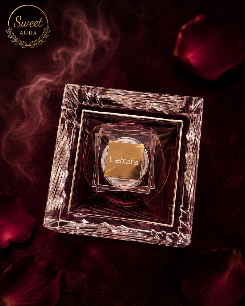 May include: A square glass perfume bottle with a faceted, crystal-like border and a gold-colored square cap featuring the text Lattafa. The bottle contains a deep red liquid, and the top left corner displays the text Sweet AURA in a circle.