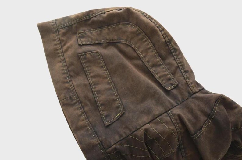 Puede incluir: Primer plano de una chaqueta con capucha marr&oacute;n. La chaqueta presenta dos bolsillos rectangulares y una capucha. Las costuras son visibles y la tela tiene un aspecto ligeramente desgastado. La chaqueta est&aacute; hecha de un material duradero.