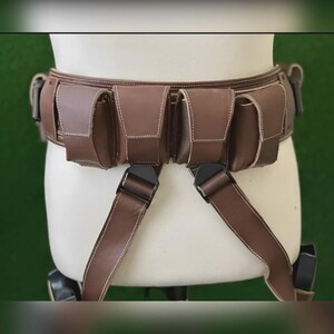 May include: A brown leather utility belt with multiple pouches and adjustable straps. The belt features white stitching and black buckles. The pouches are rectangular and appear to be designed for carrying small items. The belt is displayed on a mannequin.