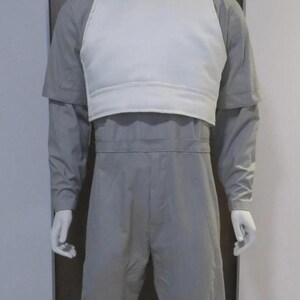 May include: A mannequin wearing a grey jumpsuit with a white vest. The jumpsuit has long sleeves and a high collar. The white vest is worn over the jumpsuit and has a V-neck design.