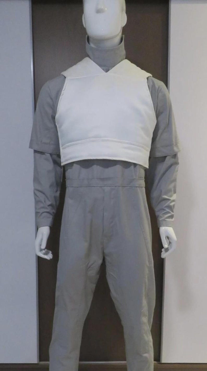 May include: A mannequin wearing a grey jumpsuit with a white vest. The jumpsuit has long sleeves and a high collar. The white vest is worn over the jumpsuit and has a V-neck design.
