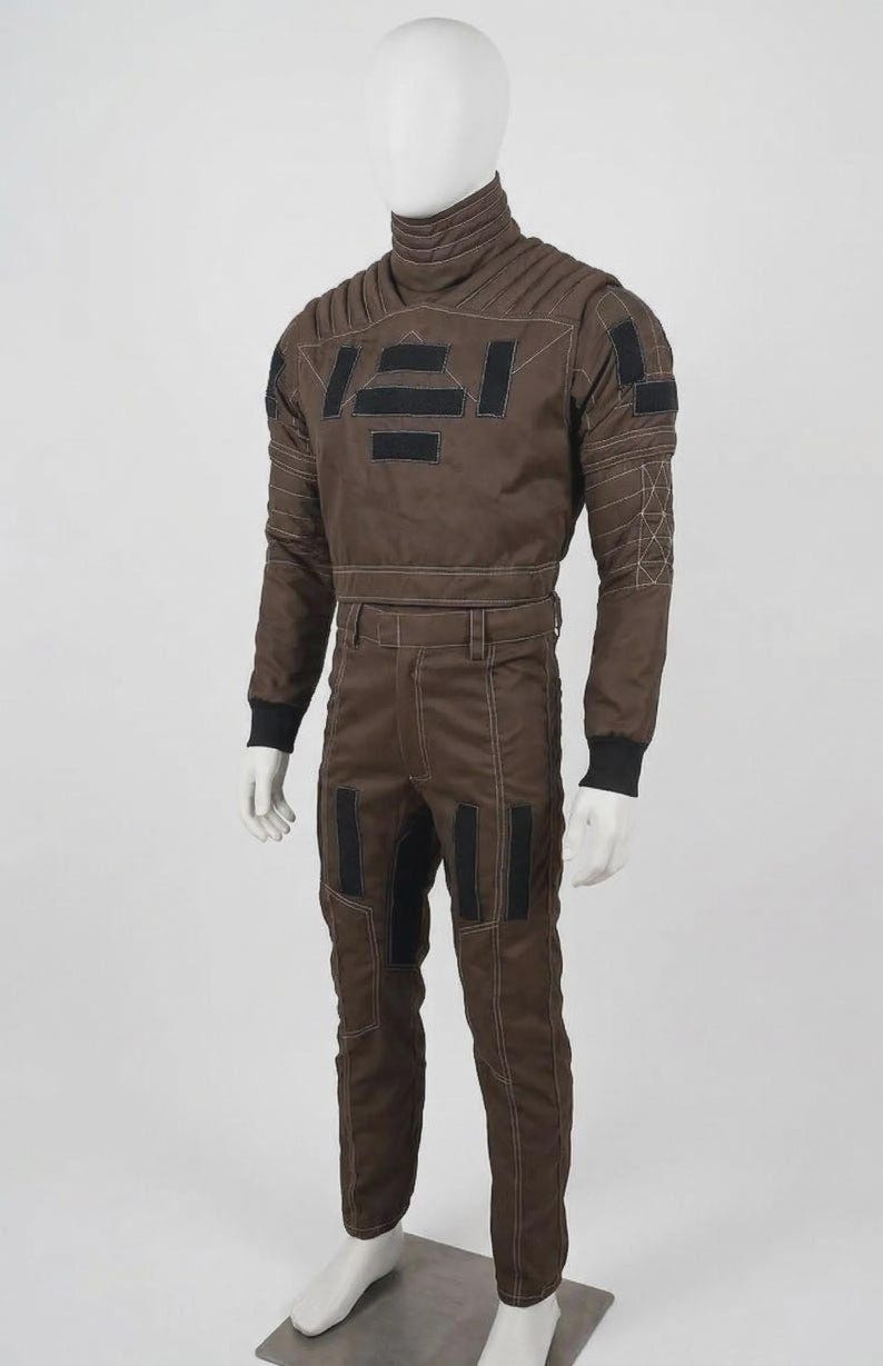 May include: A brown jumpsuit with a high collar, long sleeves, and matching pants. The suit features black rectangular patches and white stitching details. The mannequin is standing on a metal base.