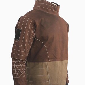 May include: A brown jacket featuring a high collar, long sleeves with white geometric stitching, and a rectangular black patch on the upper arm. The torso is divided into a dark brown upper section and a lighter brown lower section with horizontal stitching.