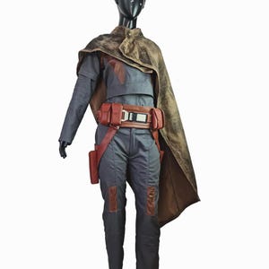 May include: A dark gray jumpsuit with long sleeves and matching trousers, featuring brown leather-like knee patches. A brown utility belt with pouches and a central buckle is worn at the waist, paired with a distressed brown cape draped over one shoulder.