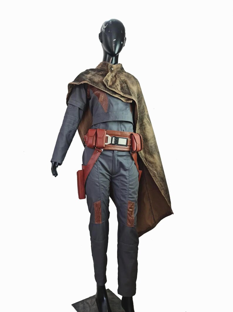 May include: A dark gray jumpsuit with long sleeves and matching trousers, featuring brown leather-like knee patches. A brown utility belt with pouches and a central buckle is worn at the waist, paired with a distressed brown cape draped over one shoulder.
