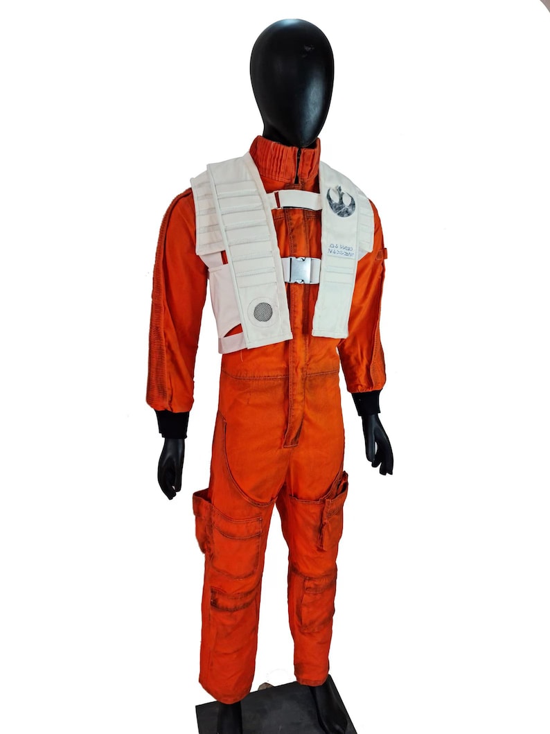 May include: A bright orange flight jumpsuit with a high collar and black cuffs, worn with a white tactical vest featuring ribbed panels, a silver buckle closure, and a circular emblem on the chest. The suit includes multiple utility pockets on the legs.
