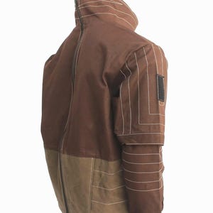 May include: A brown jacket featuring a high collar, a full-length back zipper, and a lighter brown lower panel. The sleeves are detailed with horizontal white topstitching and a black rectangular patch on the upper arm, finished with black cuffs.