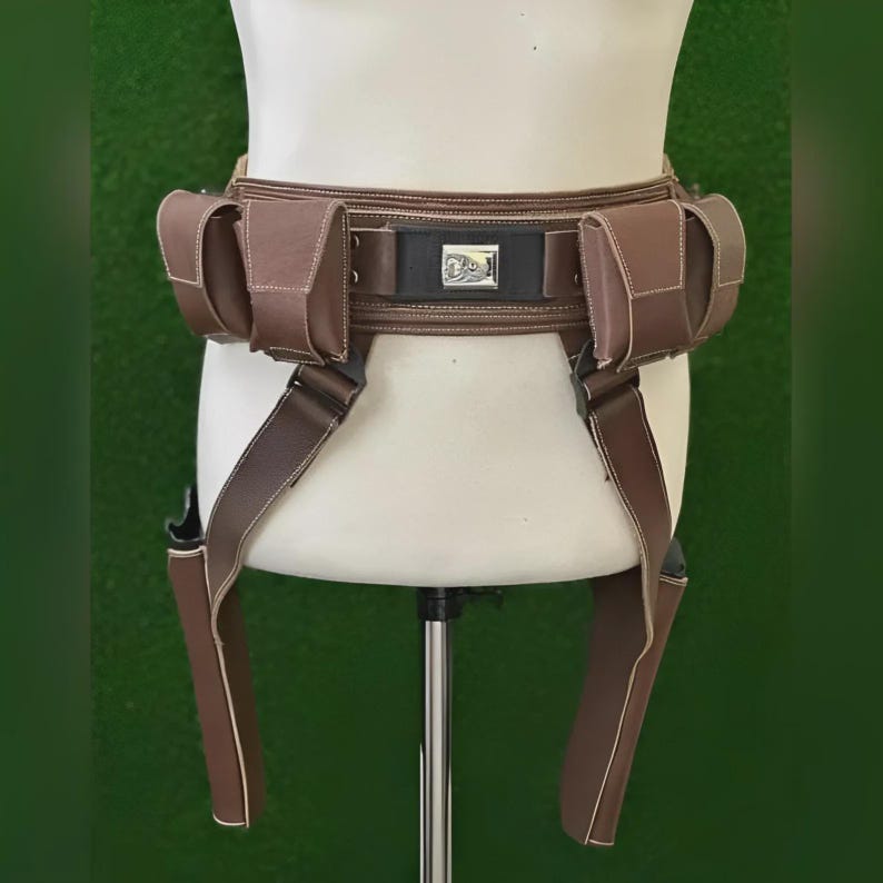 May include: Brown leather utility belt with pouches and suspenders. The belt features a central rectangular buckle with a silver emblem. The pouches are rectangular and stitched with white thread. The suspenders are attached to the belt and hang down.