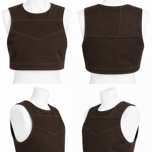 May include: A brown vest displayed on a white mannequin. The vest has a rounded neckline and a series of vertical stitched lines on the shoulders. A decorative stitched design is on the front. The vest is made of a sturdy material.