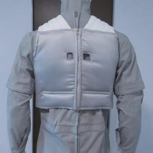 May include: A light grey jumpsuit with a matching vest and high collar, displayed on a mannequin. The vest has a zip closure and two square cutouts. The outfit appears to be made of a smooth, possibly synthetic, fabric.