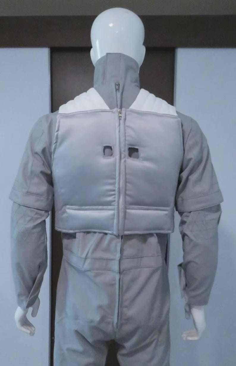 May include: A light grey jumpsuit with a matching vest and high collar, displayed on a mannequin. The vest has a zip closure and two square cutouts. The outfit appears to be made of a smooth, possibly synthetic, fabric.