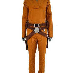 May include: A mustard yellow long-sleeved jumpsuit with a V-neckline, paired with a brown utility belt featuring a central metallic hexagonal buckle and two side holsters. The garment has a structured fit with visible vertical seam detailing.
