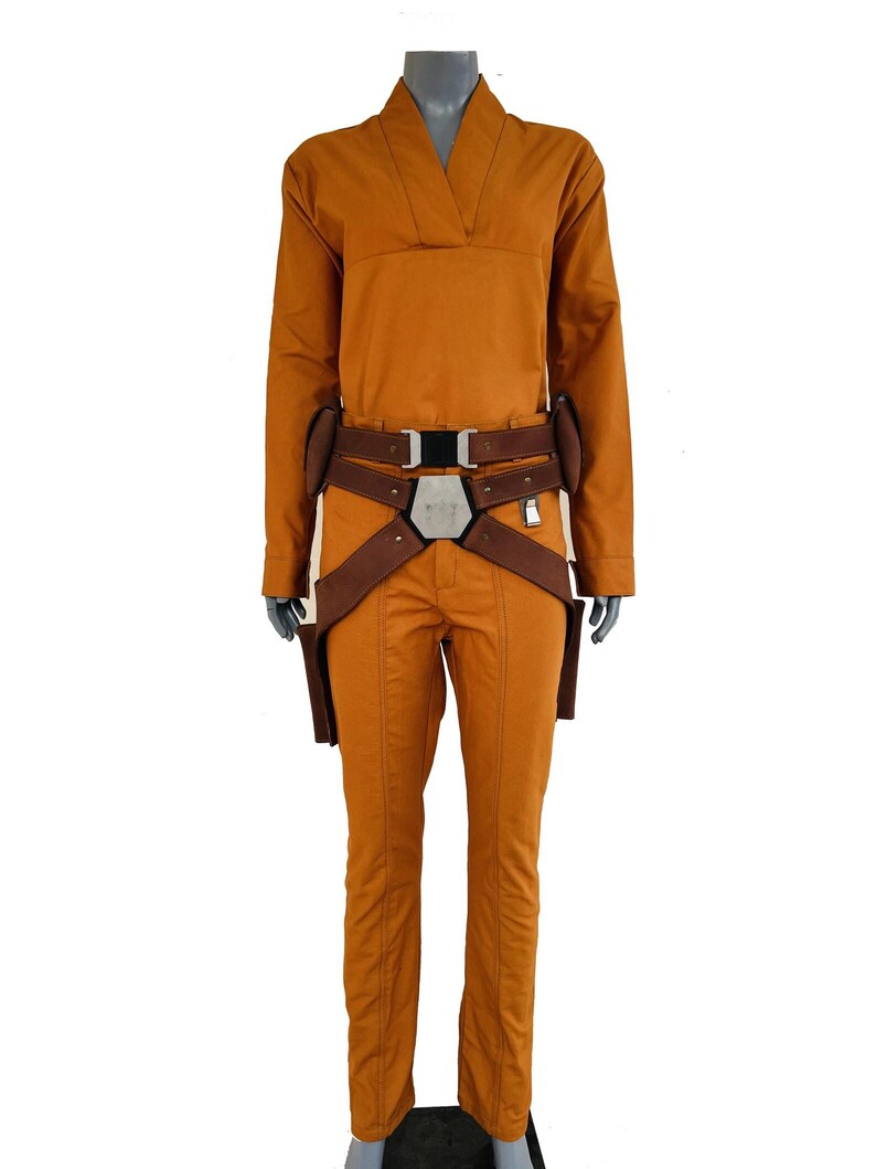 May include: A mustard yellow long-sleeved jumpsuit with a V-neckline, paired with a brown utility belt featuring a central metallic hexagonal buckle and two side holsters. The garment has a structured fit with visible vertical seam detailing.