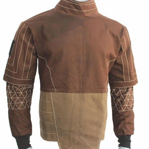 May include: A brown long-sleeve shirt featuring a high collar, a lighter brown lower panel, and white topstitching. The sleeves have a geometric triangular pattern on the forearms, while the cuffs are black. The garment has a structured design.