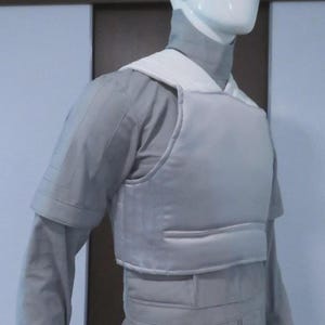 May include: A light grey protective vest and uniform on a mannequin. The vest has a white collar and is worn over a long-sleeved shirt and trousers. The outfit appears to be made of a durable material.
