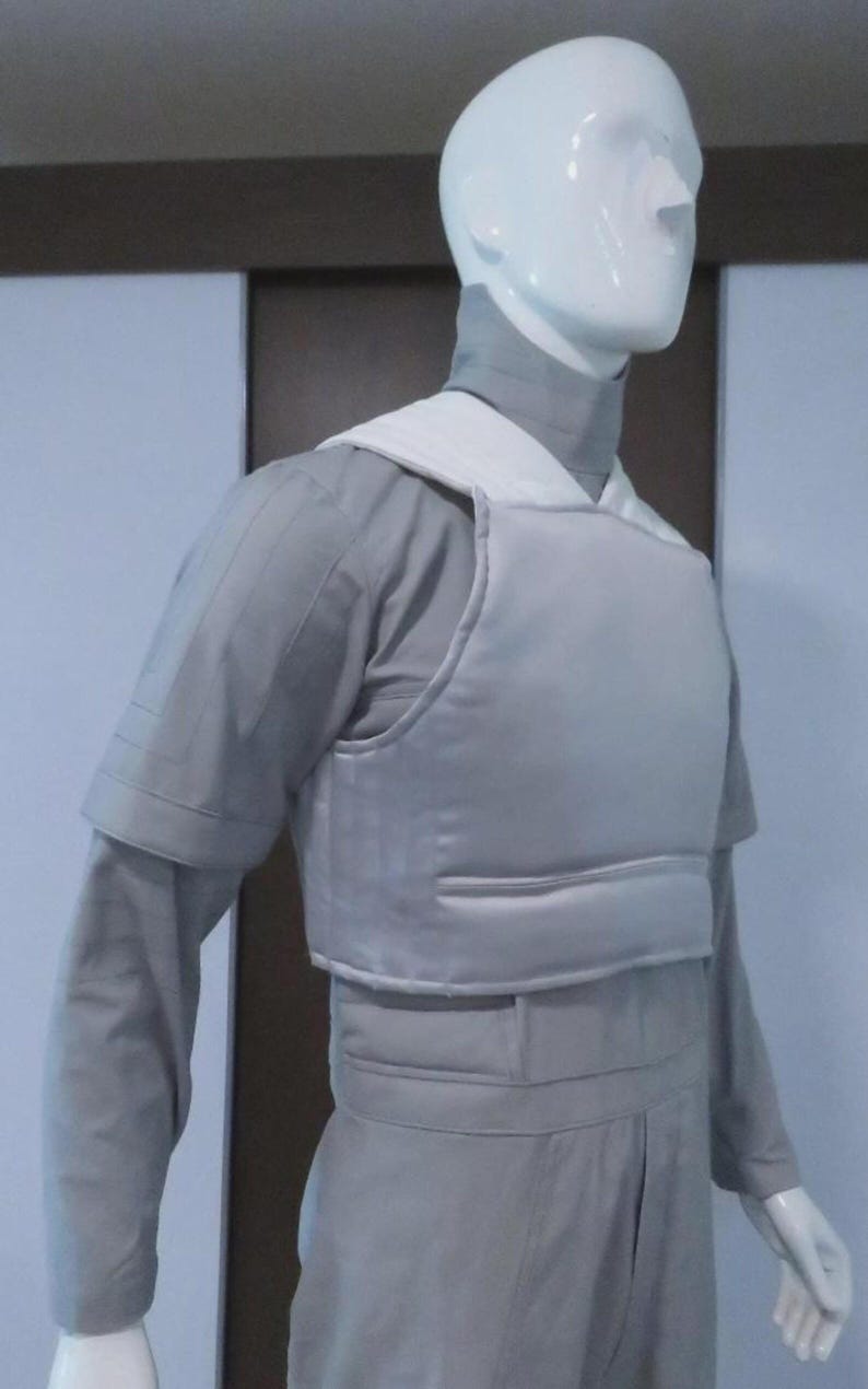 May include: A light grey protective vest and uniform on a mannequin. The vest has a white collar and is worn over a long-sleeved shirt and trousers. The outfit appears to be made of a durable material.