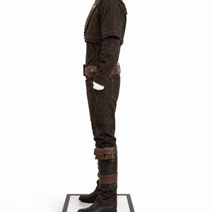 May include: A brown costume displayed on a white mannequin. The outfit includes a jacket, pants, belt, and boots. The jacket has a high collar and decorative stitching. The pants have pockets and the boots are black. The costume is displayed on a black and white platform.