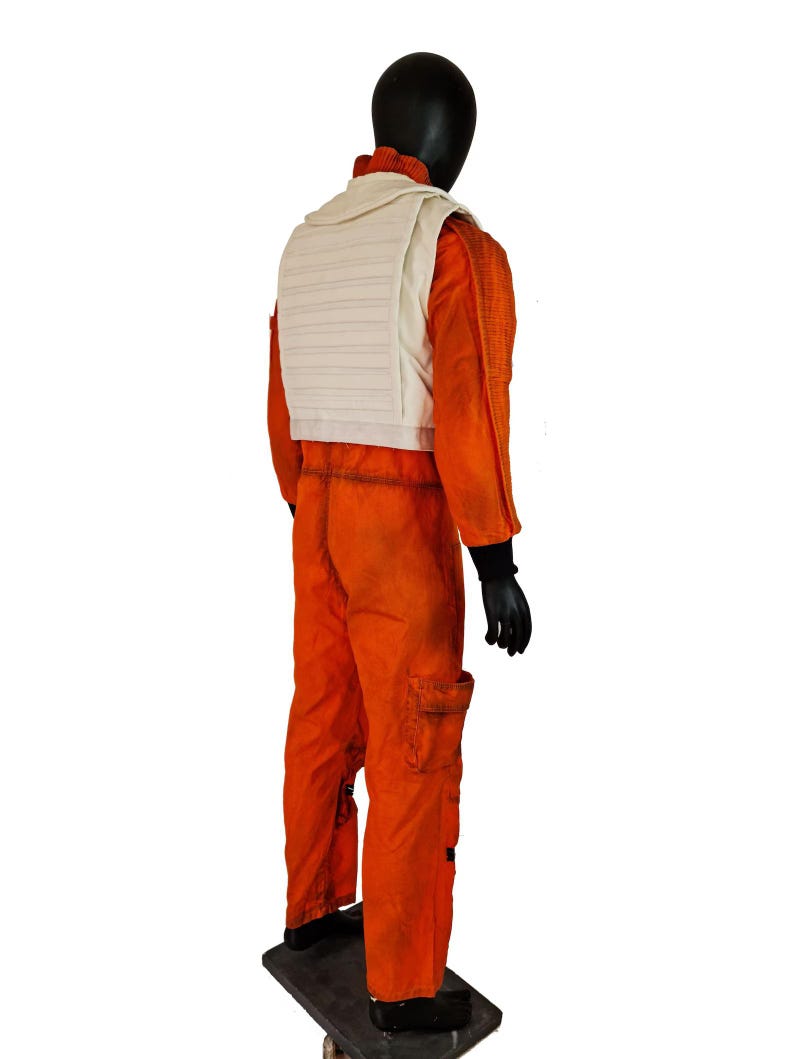 May include: A bright orange flight jumpsuit featuring a textured fabric, long sleeves with black cuffs, and a white quilted vest worn over the top. The jumpsuit includes a cargo pocket on the leg and a high collar, displayed on a black mannequin.