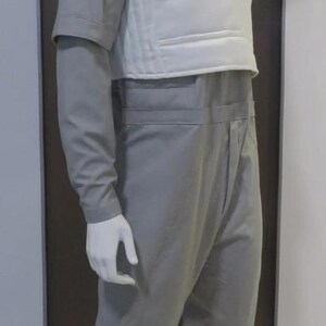 May include: A mannequin wearing a grey jumpsuit and a white vest. The jumpsuit has long sleeves and a high collar. The vest is padded and covers the chest and upper back. The mannequin is standing on a round base.