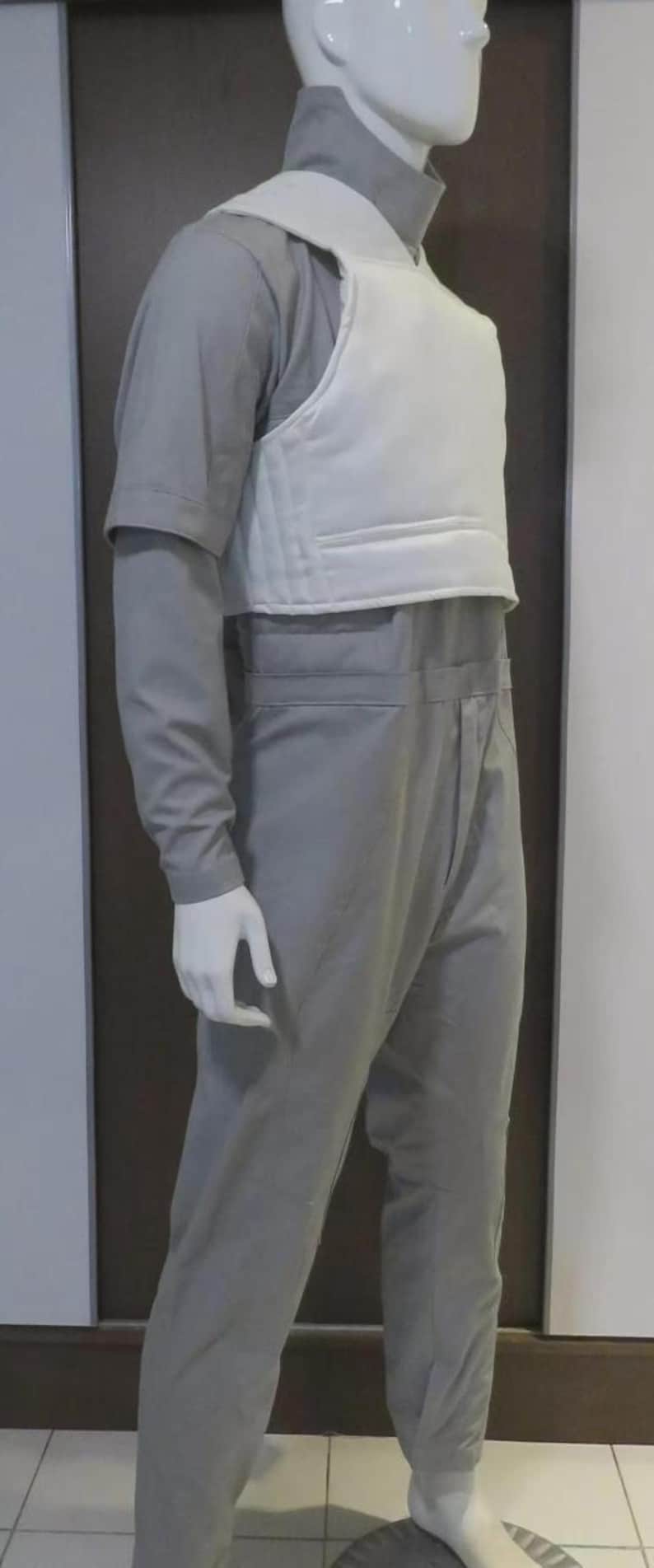 May include: A mannequin wearing a grey jumpsuit and a white vest. The jumpsuit has long sleeves and a high collar. The vest is padded and covers the chest and upper back. The mannequin is standing on a round base.