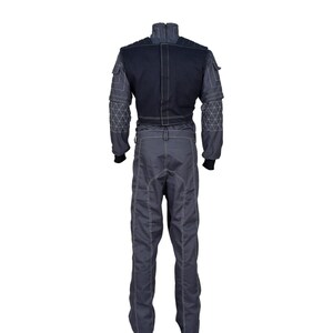 May include: A gray racing suit with a black upper section. The suit features white stitching, geometric patterns on the sleeves, and black cuffs. It has a high collar and multiple pockets, designed for professional racing.