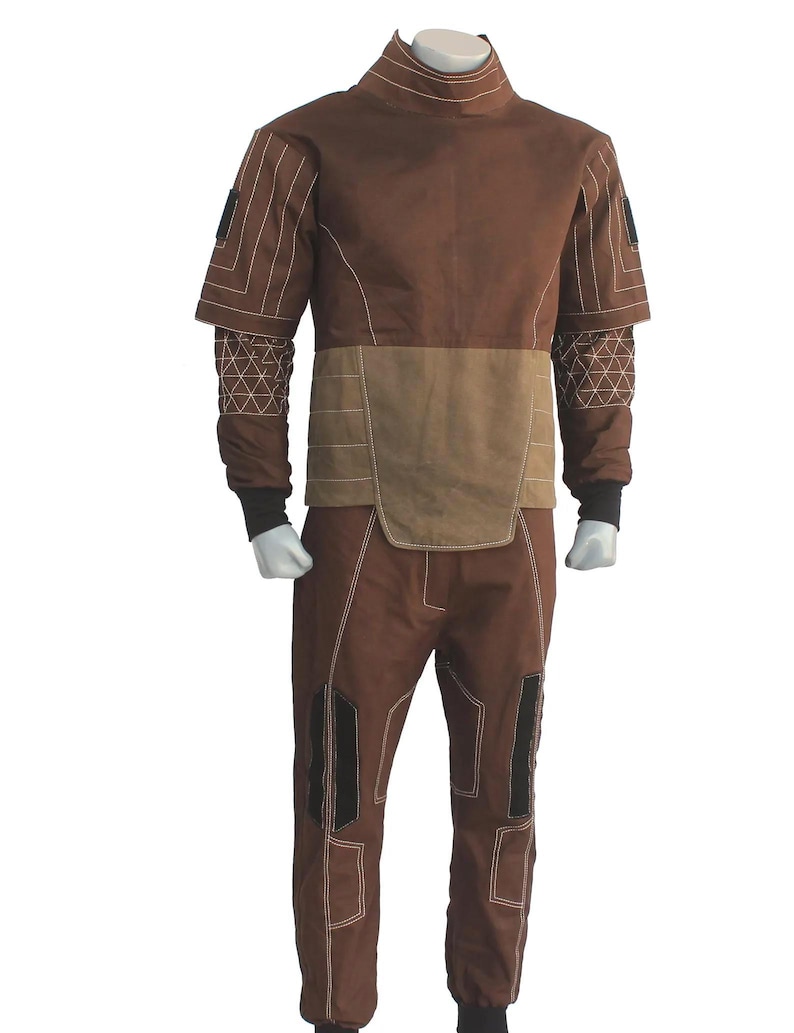 May include: A brown two-piece outfit consisting of a high-neck short-sleeve top with a lighter brown midsection and matching trousers. The garment features white contrast stitching, black sleeve cuffs, and black rectangular accents on the sleeves and legs.