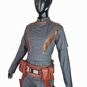 May include: A dark gray long-sleeved jumpsuit featuring a high collar, brown textured panels along the torso, and horizontal stitching on the sleeves. A brown utility belt with multiple pouches and a central metallic buckle is worn at the waist.