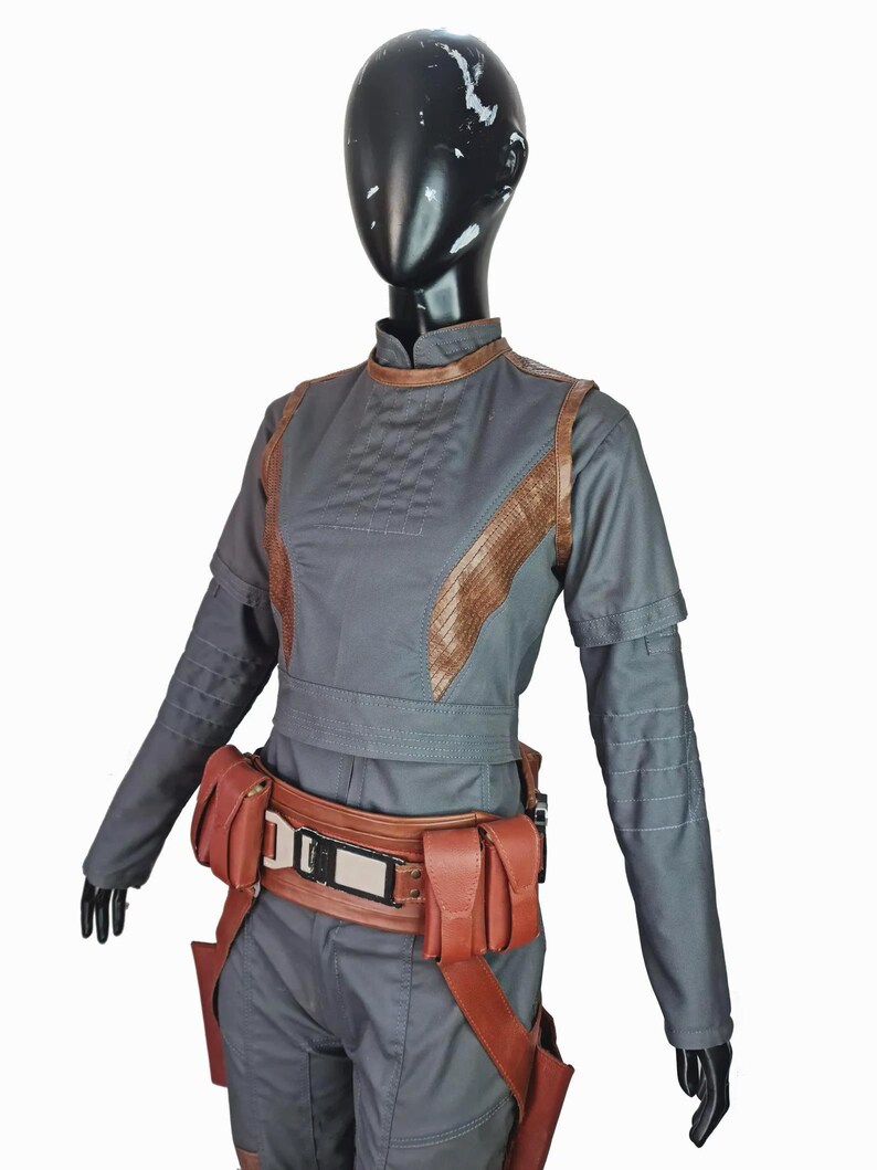 May include: A dark gray long-sleeved jumpsuit featuring a high collar, brown textured panels along the torso, and horizontal stitching on the sleeves. A brown utility belt with multiple pouches and a central metallic buckle is worn at the waist.