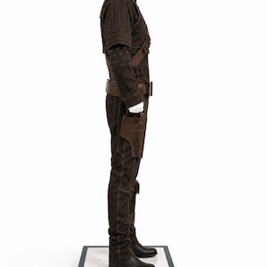 May include: A brown costume with a high collar, shoulder straps, and a belt. The outfit includes a jacket, pants, and boots. A holster is attached to the belt. The costume is displayed on a white mannequin.