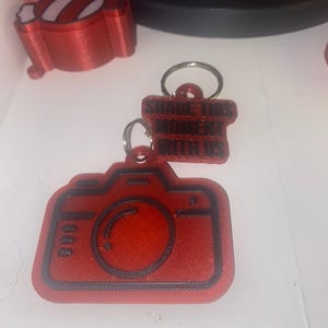 May include: A red camera-shaped keychain with black detailing and a smaller rectangular tag attached by a metal ring. The tag features the black embossed text "SHARE THIS MOMENT WITH US" in a bold, sans-serif font, creating a clear visual contrast.
