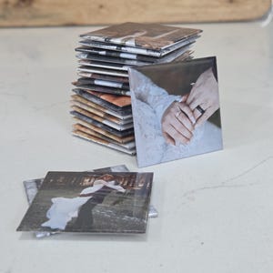 Pode incluir: Uma pilha de porta-copos quadrados com impress&otilde;es fotogr&aacute;ficas. O porta-copos do topo exibe um close-up de duas m&atilde;os com alian&ccedil;as, enquanto o porta-copos em primeiro plano mostra um casal em trajes formais abra&ccedil;ado em um ambiente florestal.