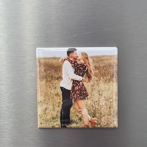 Pode incluir: Um &iacute;man quadrado para frigor&iacute;fico com uma fotografia de duas pessoas a abra&ccedil;arem-se e a beijarem-se num campo de erva. A pessoa &agrave; esquerda veste uma camisa branca de manga comprida e cal&ccedil;as escuras, a da direita um vestido floral.