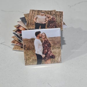 Pode incluir: Uma pilha de porta-copos quadrados com impress&otilde;es fotogr&aacute;ficas personalizadas. O porta-copos superior mostra um casal num campo, com uma pessoa a beijar a outra na bochecha. O casal veste roupa casual, incluindo uma camisa clara e cal&ccedil;as escuras.