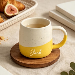 Pode incluir: Uma caneca de cerâmica branca e amarela com o nome "Jack" em itálico, sobre um descanso de madeira. A caneca contém um líquido transparente. Uma cesta com croissants e um figo está ao fundo.
