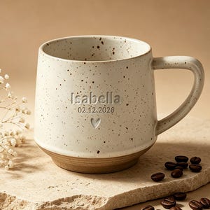 Puede incluir: Una taza de cerámica de color blanco roto con motas, que presenta el nombre Isabella, la fecha 02.12.2026 y un pequeño corazón grabado en el frente. La taza tiene un asa lisa y una base marrón sólida, acompañada de flores blancas secas y granos de café.