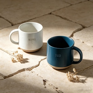 Puede incluir: Dos tazas de cerámica, una blanca y otra azul, con bases marrones. La taza blanca tiene la palabra "MAMA" y un corazón, mientras que la taza azul dice "DAD" con un corazón. Ambas tazas tienen una textura moteada.