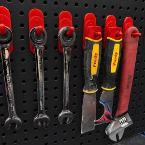May include: Three chrome ratcheting wrenches, two putty knives with yellow and black handles labeled Purdy, and one adjustable wrench with a red handle, all hanging on red plastic hooks attached to a black perforated metal pegboard panel.