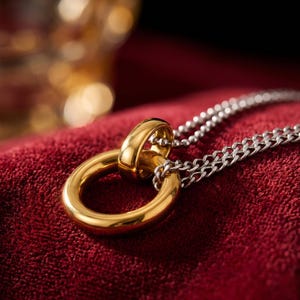 Può includere: Una collana con pendente dorato con un grande anello circolare e un anello più piccolo, collegati a una catena d'argento. Il gioiello è appoggiato su una superficie di velluto rosso, con uno sfondo sfocato.