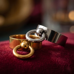 May include: Two metal rings, one gold-toned and one silver-toned, each featuring a smooth, flat band with a small, circular charm attached to the front. The gold ring is positioned slightly in front of the silver ring, both displaying a polished finish.