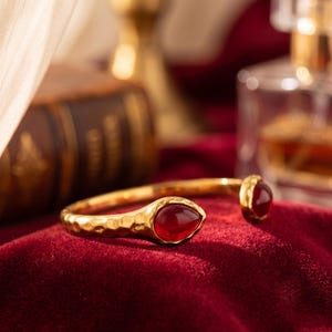 May include: A gold-toned open cuff bracelet featuring a hammered metal texture and two teardrop-shaped red gemstones at each end. The bracelet has a polished finish and a flexible design that allows for an adjustable fit around the wrist.