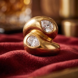 May include: A gold ring with two pear-shaped diamonds. The ring has a smooth, polished surface and is set against a red velvet background. The diamonds sparkle, reflecting light. The ring's design is modern and elegant, suitable for a special occasion.
