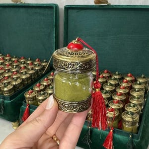 May include: A small glass jar with a decorative gold lid and a red tassel, filled with green powder, held in front of a green velvet box. The jar has ornate detailing. Other jars are visible in the background.