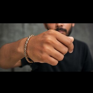 May include: A gold-coloured, braided bracelet. The bracelet is worn on a person's wrist, with the hand in focus. The bracelet has a twisted rope design. The background is a blurred grey wall.
